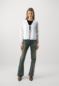 Cardigan en dentelle blanche à manches longues, devant ouvert et détail à lacets. Associé à un jean bleu délavé évasé et des bottes beiges à bout pointu.