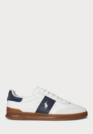 White low-top sneaker with navy side panel and Polo logo, brown rubber sole, white laces, and navy suede heel accent.