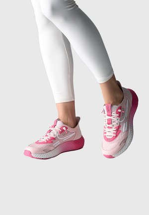 Person trägt weiße Leggings und rosa Laufschuhe mit strukturierten Sohlen, steht vor einem schlichten hellen Hintergrund.