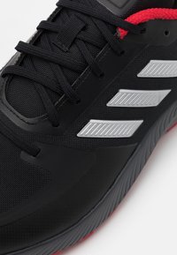 Schwarzer Sport Schuh mit schwarzen Schnürsenkeln, drei silbernen Streifen an der Seite, roten Stoffakzenten und grauer Sohle mit strukturiertem Profil.