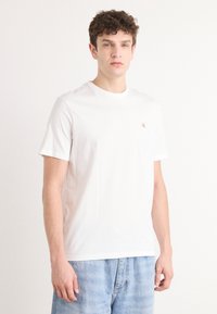 Weißes Baumwoll-T-Shirt mit Rundhalsausschnitt, kurzen Ärmeln und einem kleinen roten Logo auf der linken Brust. Modell trägt hellblaue Denim-Shorts.