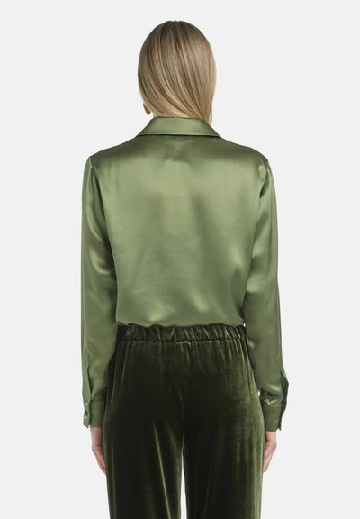 Zelena satenasta bluza dugih rukava s okovratnikom; stražnji pogled prikazuje glatku teksturu tkanine. Uparen s tamnozelenim baršunastim hlačama.