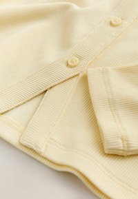 Tessuto giallo a coste con strisce testurizzate e finitura morbida. Presenta bottoni rotondi di colore crema e dettaglio della manica pieghettata.