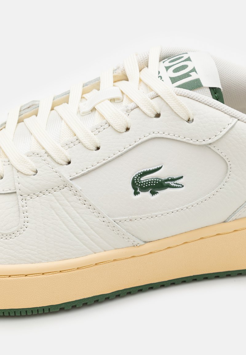 Baskets en cuir blanc avec une surface texturée, dotées d'un logo Lacoste vert, de lacets crème et d'une semelle en caoutchouc beige avec des motifs verts.