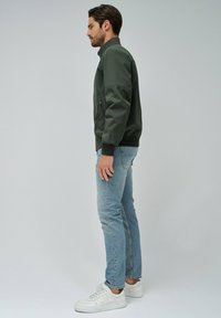 Chaqueta bomber verde oscuro con un bolsillo con cremallera, puños y cuello negros, combinada con jeans azul claro y zapatillas blancas.
