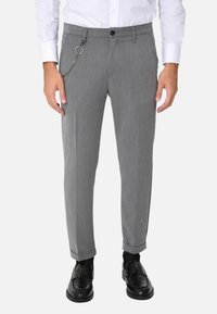 Pantaloni grigi su misura con una finitura leggermente texture, caratterizzati da un accento a catena e chiusura con bottone. Orli arrotolati e tasche laterali visibili.