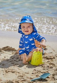 Bañador azul con patrones de cangrejos blancos y rayas naranjas, sombrero de ala ancha, sentado en la arena con un cubo verde y una pala de juguetes cerca.
