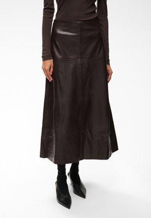 Femme portant une longue jupe en cuir marron foncé avec des chaussures noires à bout pointu et un haut à manches longues marron foncé assorti, debout sur fond blanc.