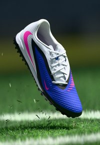 Ποδοσφαιρικό παπούτσι Nike με λευκό πάνω μέρος, μπλε και μαύρες λεπτομέρειες, ροζ σχεδιαστικά στοιχεία, υφή στην επιφάνεια και σόλα με πολλαπλές προεξοχές, τοποθετημένο σε γρασίδι.