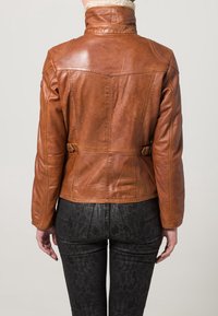 Bruine leren jas met opstaande kraag, textuurafwerking en zijkant gespaccenten, gecombineerd met zwarte legging met patroon.