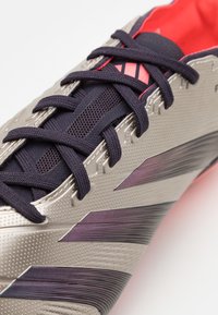 Scarpa da calcio argento e nera con superficie texturizzata, strisce viola e lacci scuri. Fodera rossa brillante visibile all'interno della scarpa.