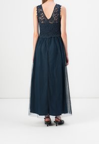 Kvinna klädd i en ärmlös marinblå spetsöverdel med genomskinlig rygg och en lång mörk tyllkjol, tillsammans med svarta låga klackskor.