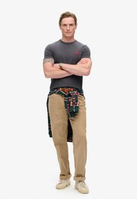 T-shirt grigio a maniche corte con logo rosso, pantaloni in corduroy color sabbia, camicia a quadri verde e rossa annodata in vita, sneakers beige.