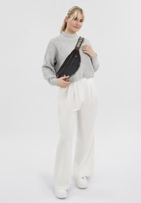 Zwarte stoffen schoudertas met twee ritscompartimenten en bruine accenten, gedragen over de tors met een gezellige grijze gebreide trui en witte broek.