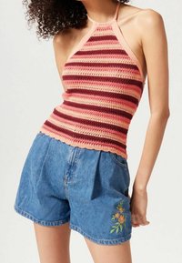 Donna che indossa un top halter senza maniche a righe rosa e rosse e pantaloni corti in denim blu a vita alta con ricami floreali sulla gamba sinistra.