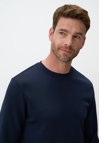 Felpa girocollo in cotone blu navy con texture liscia, maniche lunghe e dettagli sulle cuciture delle spalle, indossata da un uomo con leggera barba.