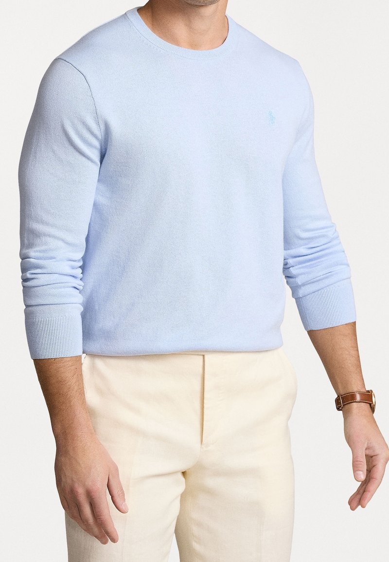 Jasnoniebieski sweter z długim rękawem, wykonany z miękkiego materiału, z ribbowanymi mankietami i małym logo na piersi, zestawiony z kremowymi spodniami.