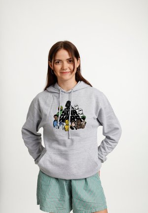 Młoda kobieta w szarej bluzie z kapturem Star Wars i zielono-paskowanych szortach, stoi z rękami w kieszeniach, uśmiechając się na białym tle.