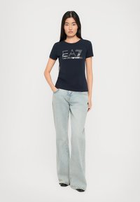 Marineblå bomull T-skjorte med et sentralt logodesign med sølvfarger, kombinert med lyse, vide jeans. Jeansen har en glatt tekstur og en tettsittende form.