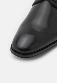 Chaussure en cuir noir avec une finition brillante, bout pointu et un détail de couture le long de la semelle. Texture lisse et design épuré.
