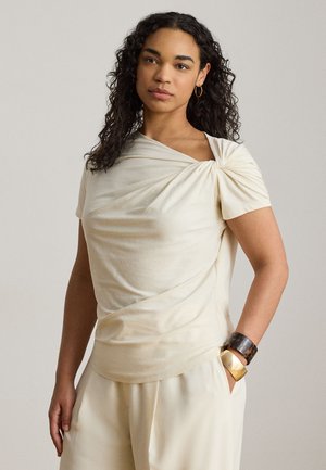 Femme aux cheveux bouclés porte un top drapé asymétrique ivoire et un pantalon ample assorti, accessoirisés de bracelets en or et en écaille.