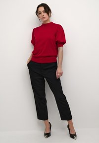 Pulover roșu cu guler înalt și mâneci bufante, asortat cu pantaloni negri pe corp și tocuri negre ascuțite. Textură netedă și siluetă ajustată.
