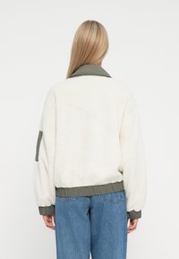 Fleece-takki vaaleassa valkoisessa, oliivinvihreällä kauluksella ja vyötäröltä, jossa on rento istuvuus ja ribbineulekämmekkeet, esittelee taskun hihassa.