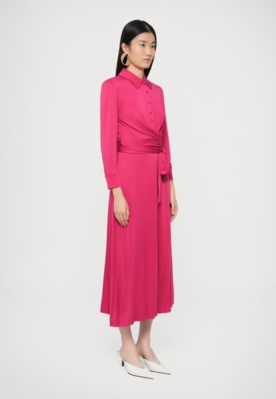 Φούξια φόρεμα τύπου φόρμας με γιακά, κουμπιά μπροστά, μακριά μανίκια και ζώνη στη μέση. Φορεμένο με λευκές ψηλοτάκουνες γόβες με μυτερή μύτη.