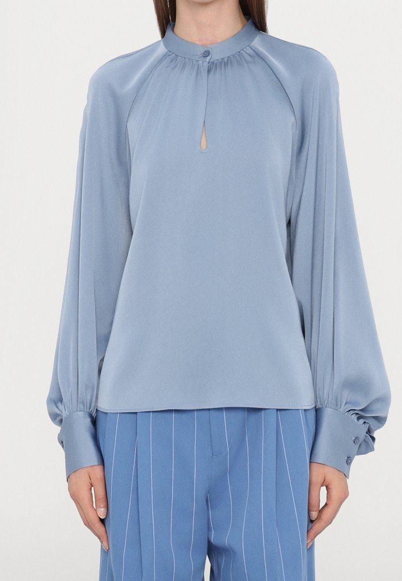Femme portant un chemisier bleu clair à manches longues avec un col en forme d'œillet boutonné et des poignets larges, associé à un pantalon bleu à fines rayures.