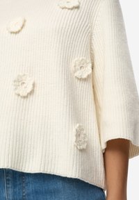 Krémový pletený svetr s žebrovanou strukturou, zdobený háčkovanými květinovými aplikacemi uspořádanými do shluků. Krátké rukávy a volný střih.