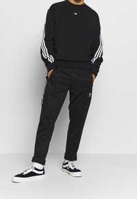 Czarna bluza z białym wzorem trzech pasków na rękawach oraz małym logo na piersi, zestawiona z czarnymi spodniami dresowymi i czarnymi sneakersami.