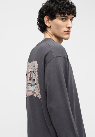 Νεαρός με σγουρά σκούρα μαλλιά που φοράει σκούρο γκρι μακρυμάνικη μπλούζα με ένα μοτίβο ορθογώνιο γραφικό στην πλάτη.