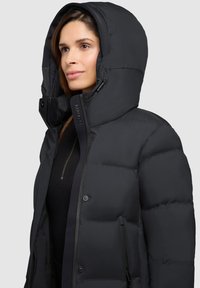 Zwarte pufferjack met een ruime capuchon, drukknoopsluiting en ritszakken. Het materiaal heeft een gladde, matte textuur met gestikte secties.