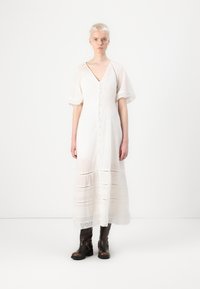 Robe maxi blanche à boutonnière avec des manches courtes évasées et des bordures en dentelle. Le tissu texturé présente des rayures horizontales. Portée avec des bottes brunes jusqu'aux genoux.