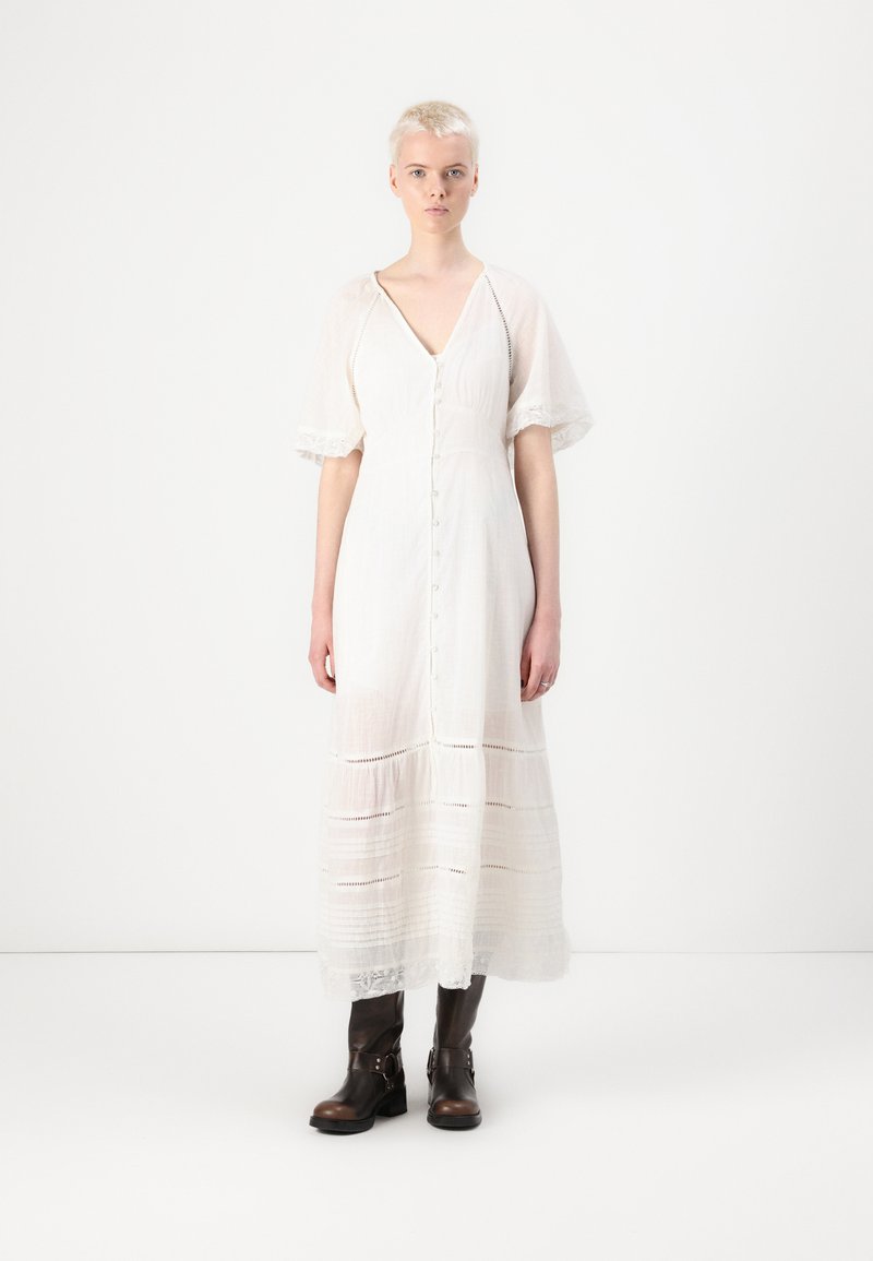 Robe maxi blanche à boutonnière avec des manches courtes évasées et des bordures en dentelle. Le tissu texturé présente des rayures horizontales. Portée avec des bottes brunes jusqu'aux genoux.