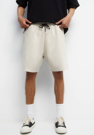 La personne porte un short beige clair avec un cordon noir, un t-shirt noir, des chaussettes blanches et des baskets noir et blanc, debout devant un fond blanc.