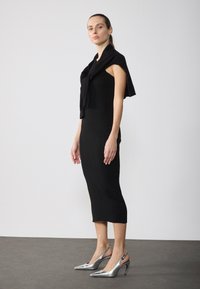 Črna rebrasta midi obleka brez rokavov, s črnim puloverjem, draperanim čez ramena. Srebrni slingback čevlji s koničastimi prsti.
