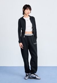 Moletão preto com fecho, top cropped branco, calças de fato de treino pretas com logótipo, ténis prateados. O material parece suave; o conjunto é casual e desportivo.