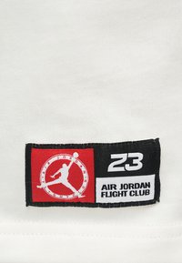 Λευκό ύφασμα με υφή. Περιέχει μια μαύρη και κόκκινη ετικέτα με τις λέξεις "AIR JORDAN FLIGHT CLUB," "23," και ένα λογότυπο σιλουέτας.