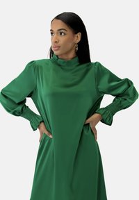 Grøn satin kjole med lange pufærmer, høj hals og rynkede manchetter. Glat tekstur og flydende silhuet.