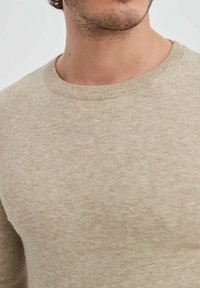 Pull en tricot beige avec un col rond, présentant une texture douce et un motif subtil, offrant une coupe décontractée.
