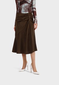 Gonna marrone, fino al ginocchio, con dettagli arricciati su un lato, realizzata in materiale morbido. Abbinata a tacchi a stiletto bianchi.