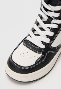 Μαύρο και άσπρο ψηλό sneaker με δερμάτινο άνω μέρος, τρυπητό μπροστινό τμήμα, λευκές ίσιες κορδόνες και αντίθετες λεπτομέρειες ραφής.