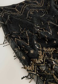 Țesătură neagră cu detalii intricate din mărgele aurii și argintii în modele geometrice; dispune de accente cu ciucuri de-a lungul marginilor.