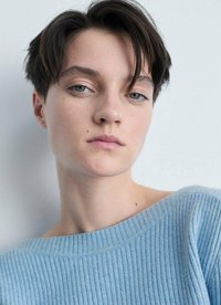 Pullover azzurro chiaro a costine con scollatura ampia, indossato da una persona con capelli corti scuri e trucco minimale, su uno sfondo neutro.