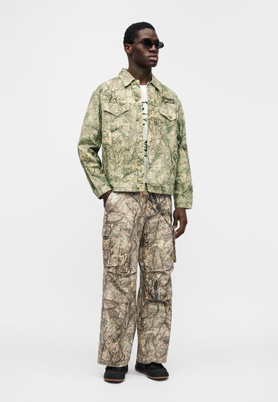 Πράσινο μπουφάν με μοτίβο καμουφλάζ και δύο τσέπες στο στήθος, συνδυασμένο με μπεζ παντελόνι καμουφλάζ τύπου cargo με πολλές τσέπες. Μαύρα παπούτσια και γυαλιά ηλίου.