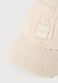 Bēšs cepure no ribota auduma ar kvadrātveida uzliku priekšpusē, kurā ir izspiedums ar zīmola nosaukumu "BOSS" vienkāršā fontā.