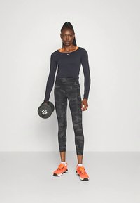 Černé sportovní tričko s dlouhým rukávem s oranžovými akcenty, kombinované s černými maskáčovými legínami. Modelka drží v jedné ruce činku a má na sobě oranžové tenisky.