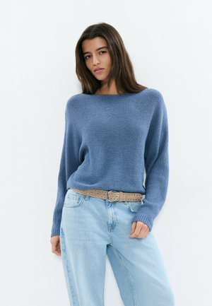 Jeune femme portant un pull bleu ample rentré dans un jean bleu clair avec une ceinture beige perforée, posant devant un fond blanc.
