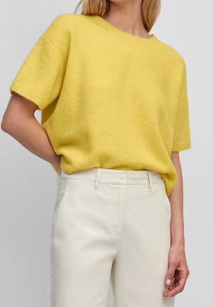 Femme portant un haut en maille jaune à manches courtes, rentré dans un pantalon blanc taille haute avec passants pour ceinture, sur un fond uni.